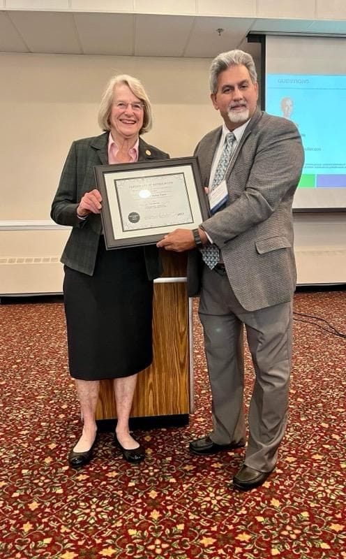 A woman in a plaid blazer and a man in a grey jacket standing in a conference hall, holding a framed VAPCP Certificate of Appreciation